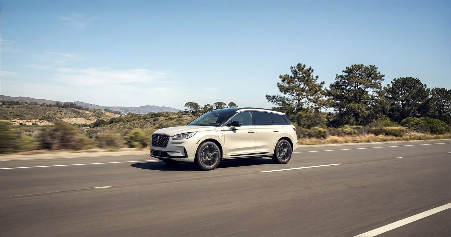 A white SUV driving on a road with pine trees and mountainous terrain in the background.