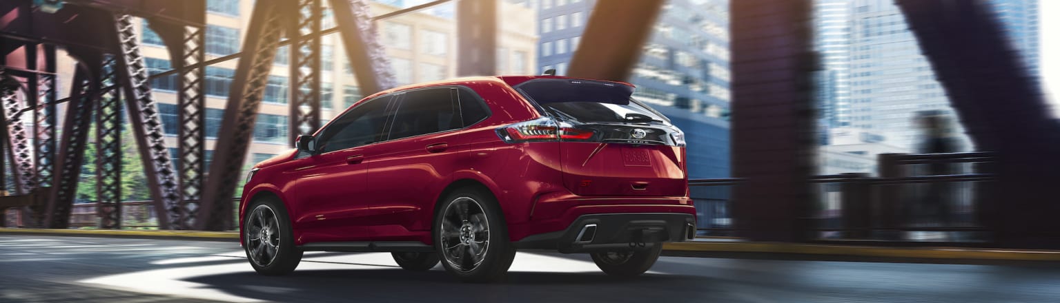 the rear end of a red ford edge suv driving on a city street with tall buildings in the background