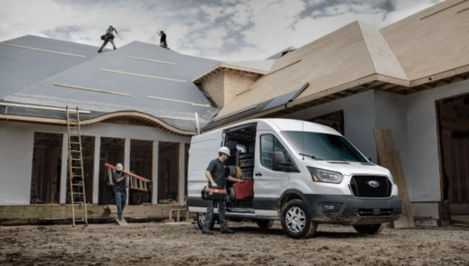A white work van parked in front of a house under construction with workers on the roof and carrying materials.