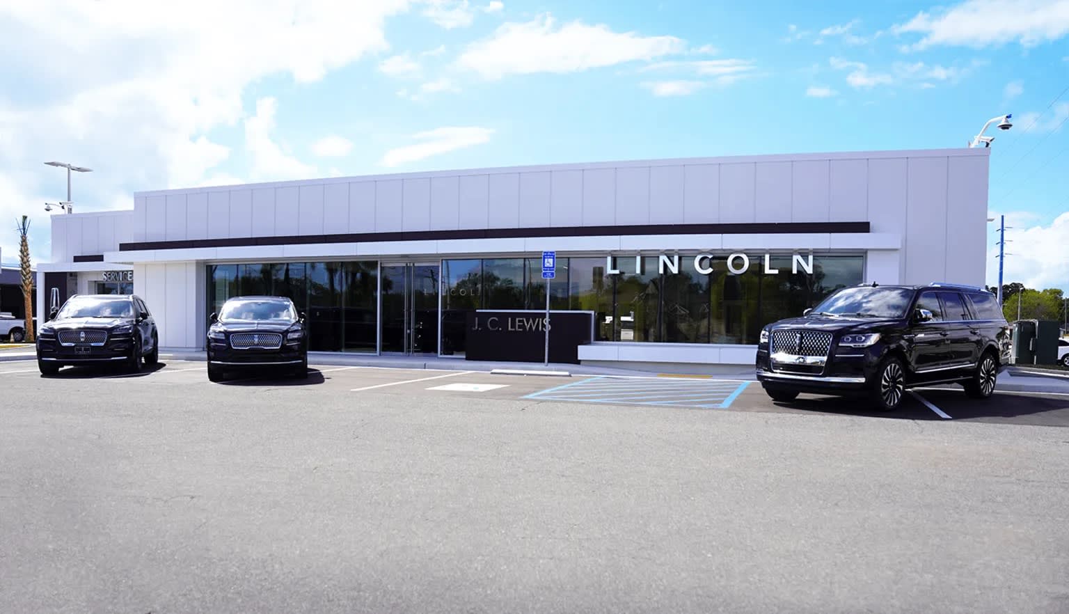 A modern, white-colored car dealership building with large glass windows, surrounded by parked vehicles in the foreground and a clear blue sky with clouds in the background.