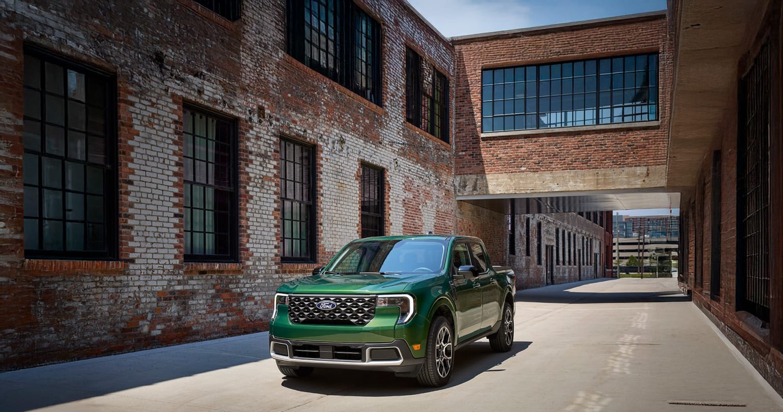 A green pickup truck parked in front of a brick building with large windows on a sunny day.