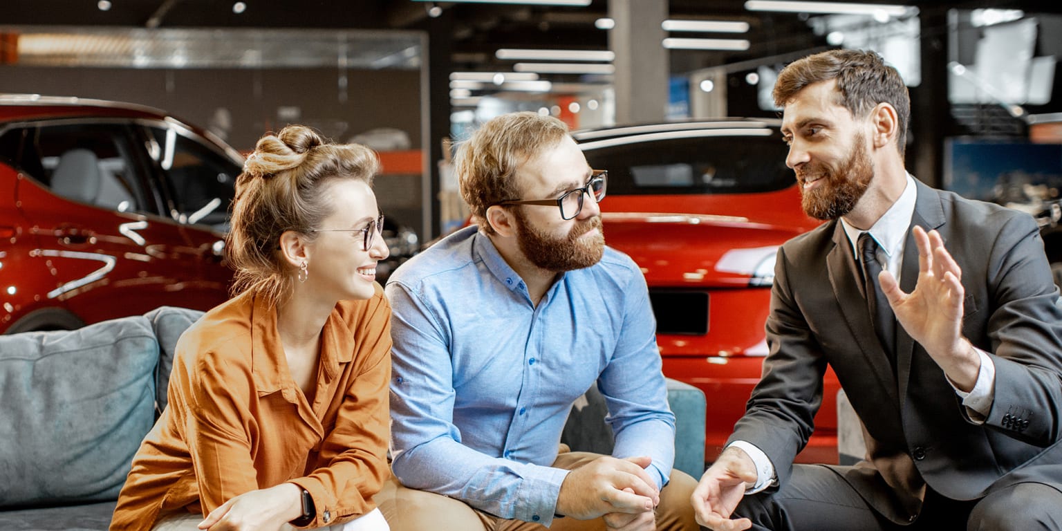 A couple sitting on a couch at a car dealership, chatting with a salesman in front of red cars.