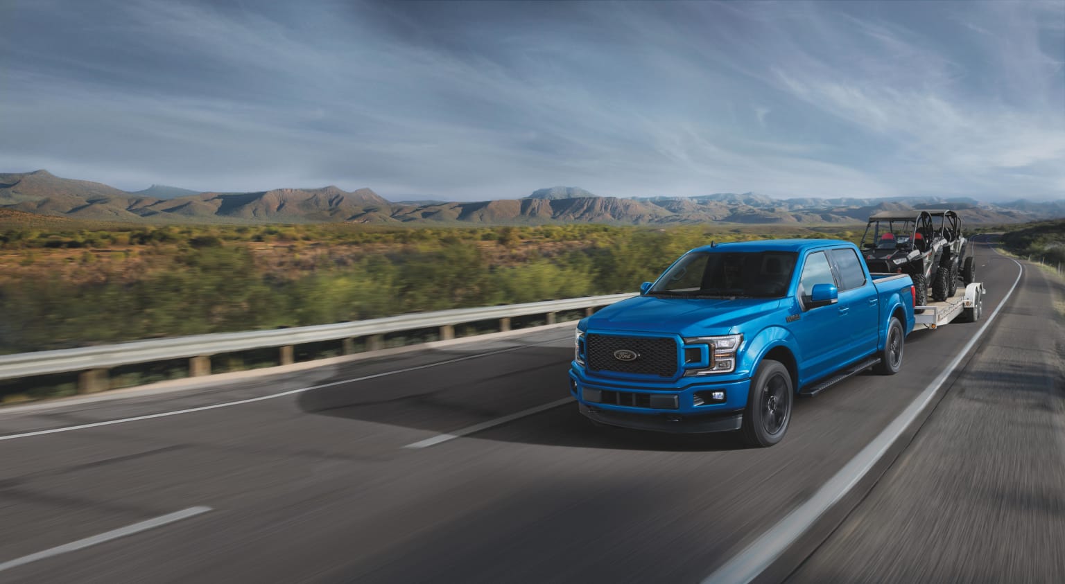 a blue truck driving down a highway next to a lush green hillside under a cloudy sky with mountains in the distance