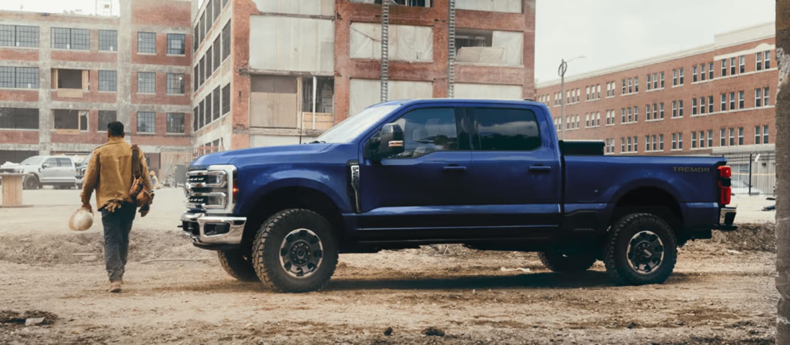 A man walking towards a blue pickup truck in a dusty lot with industrial buildings in the background.