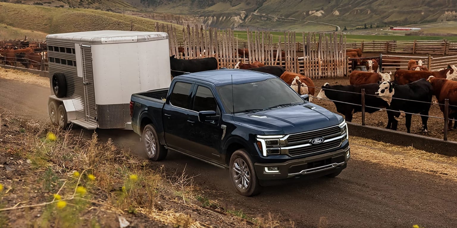 A large black pickup truck parked on a dirt road with horses in the background, in a rural landscape.