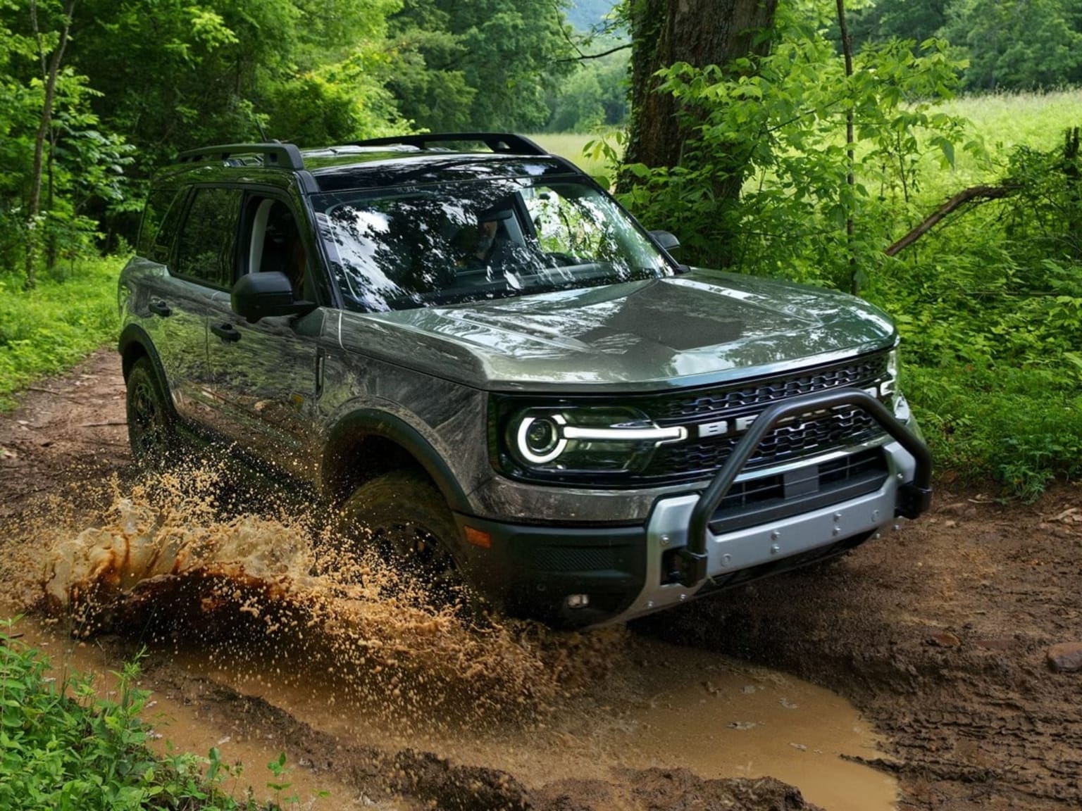 An SUV drives through a muddy puddle on a dirt road, with lush green trees and foliage in the background.