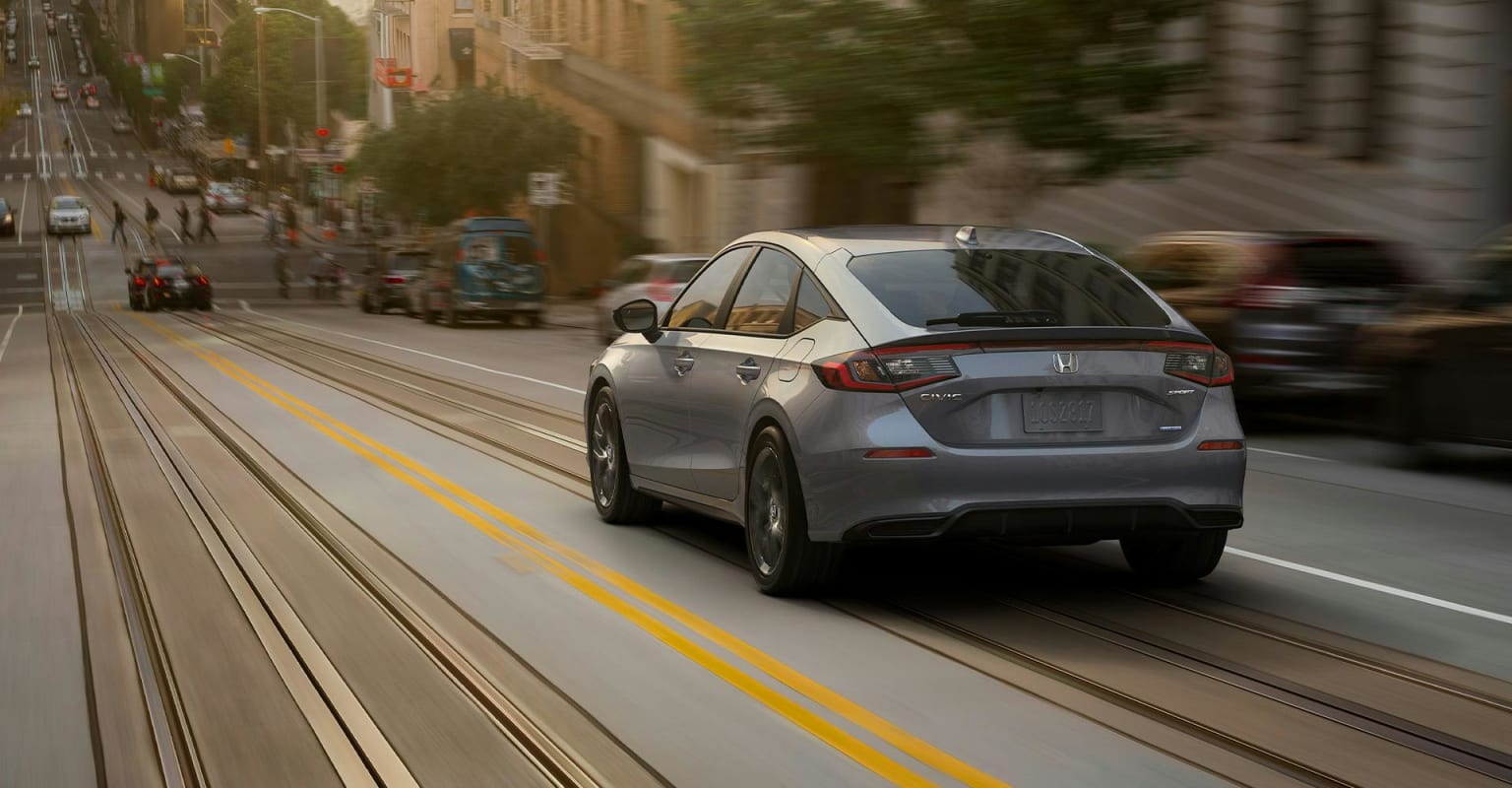A silver hatchback drives down a city street with blurred buildings and people in the background.
