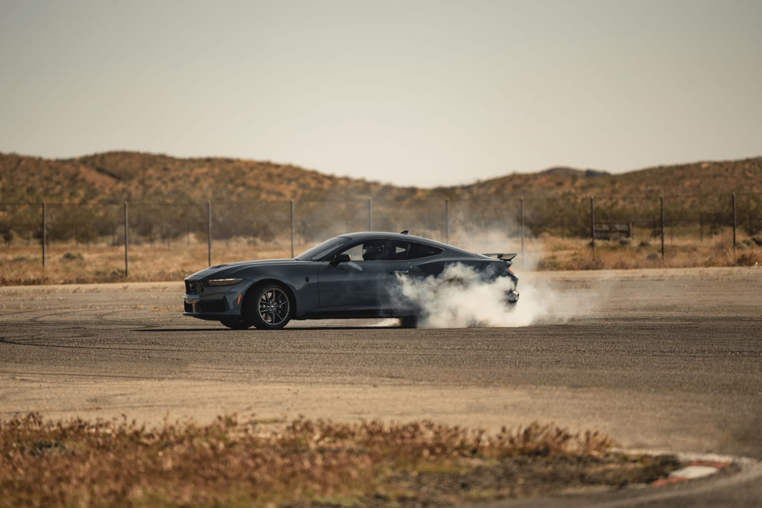 The image shows a black sports car performing a burnout on a dusty, desert-like terrain, with a mountainous background in the distance.
