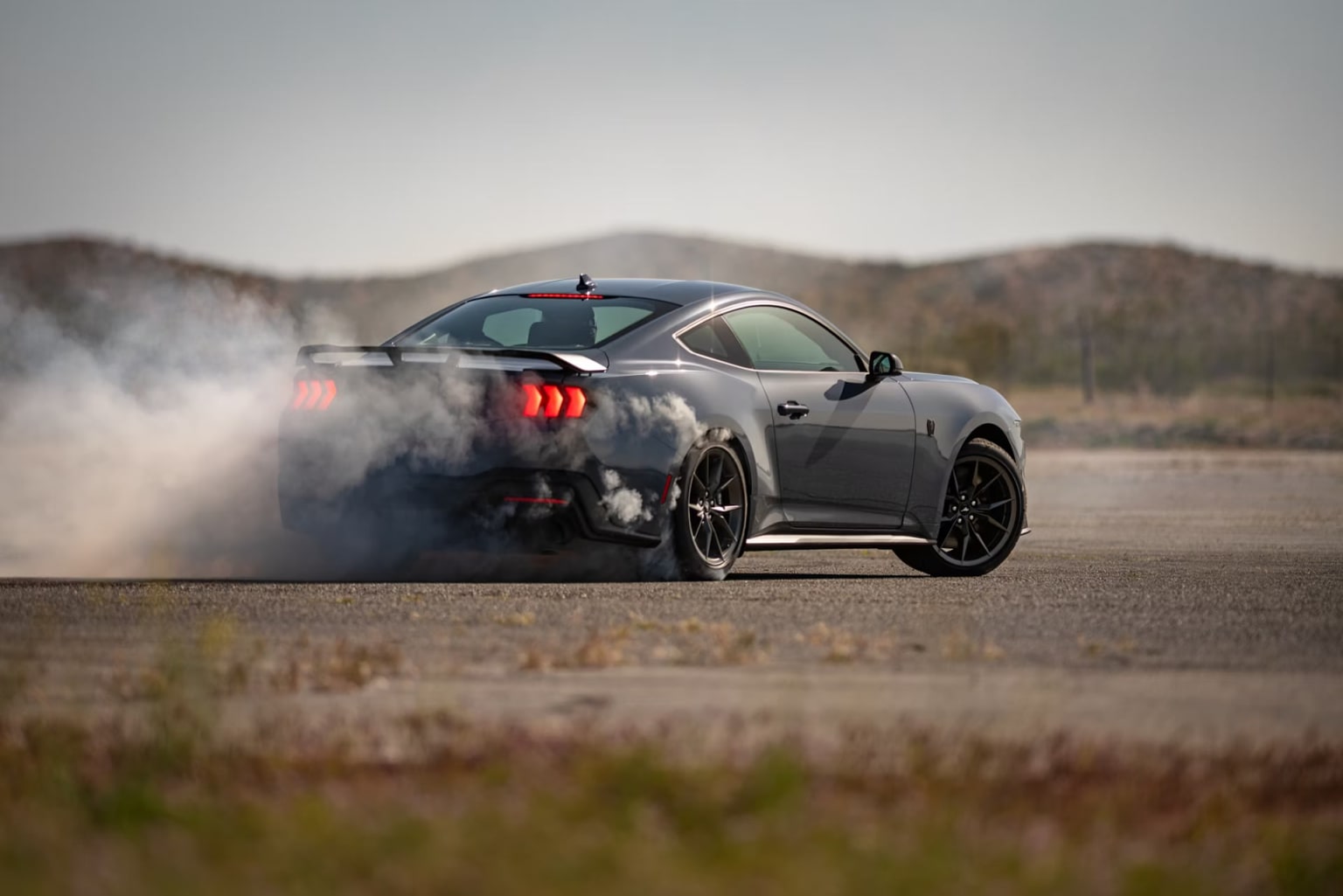 A gray sports car drifting on a dirt road with a grassy field and mountains in the background.