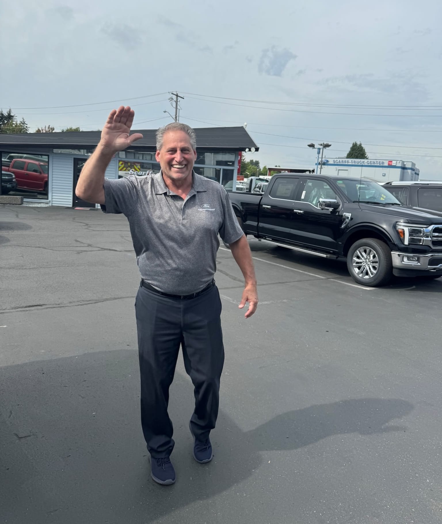 An older man in casual clothing is waving his hand in front of a car dealership with several vehicles parked outside.