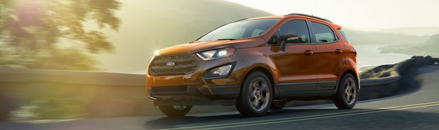 an orange suv driving down a road next to a mountain range in the sun with trees in the background