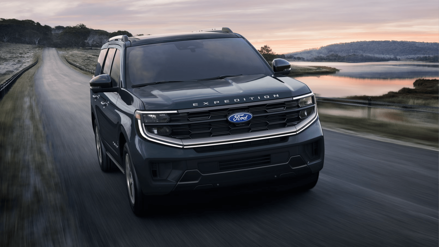 A dark green large SUV drives along a winding road beside a calm lake under a soft sunset sky.