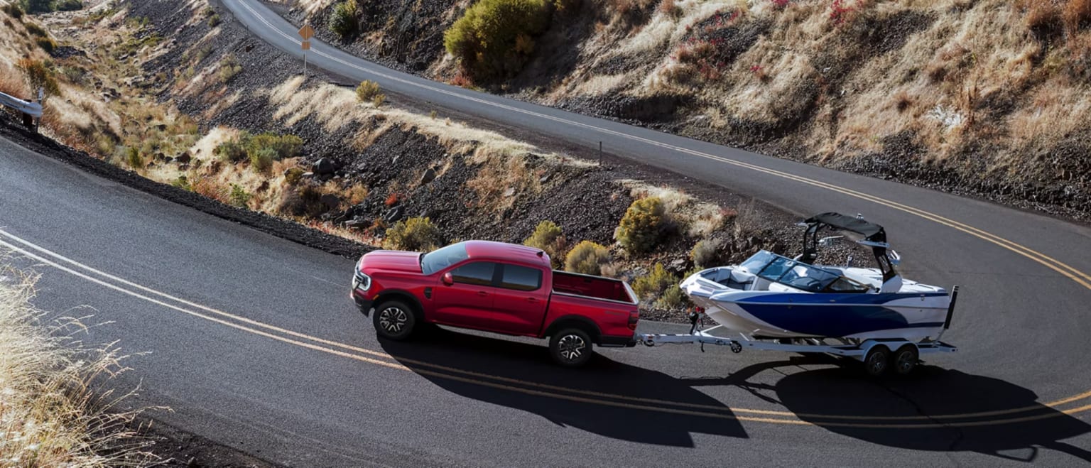 A red pickup truck towing a boat trailer on a scenic asphalt road in a dry, hilly landscape.