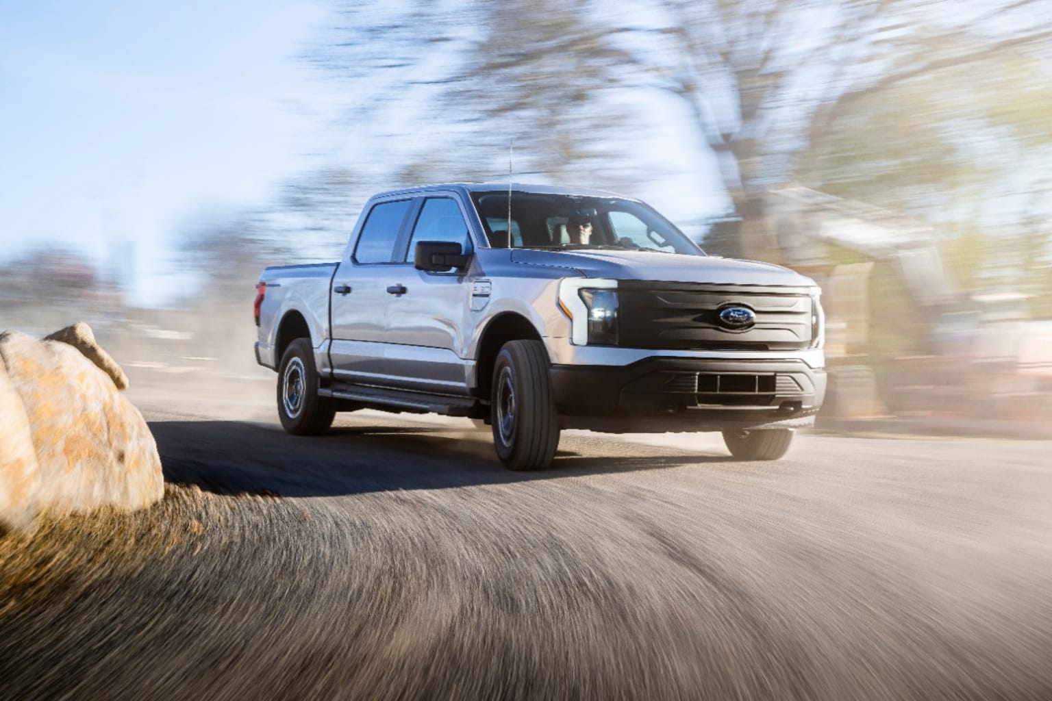 a white truck driving down a road next to a large rock in the middle of the road with trees in the background