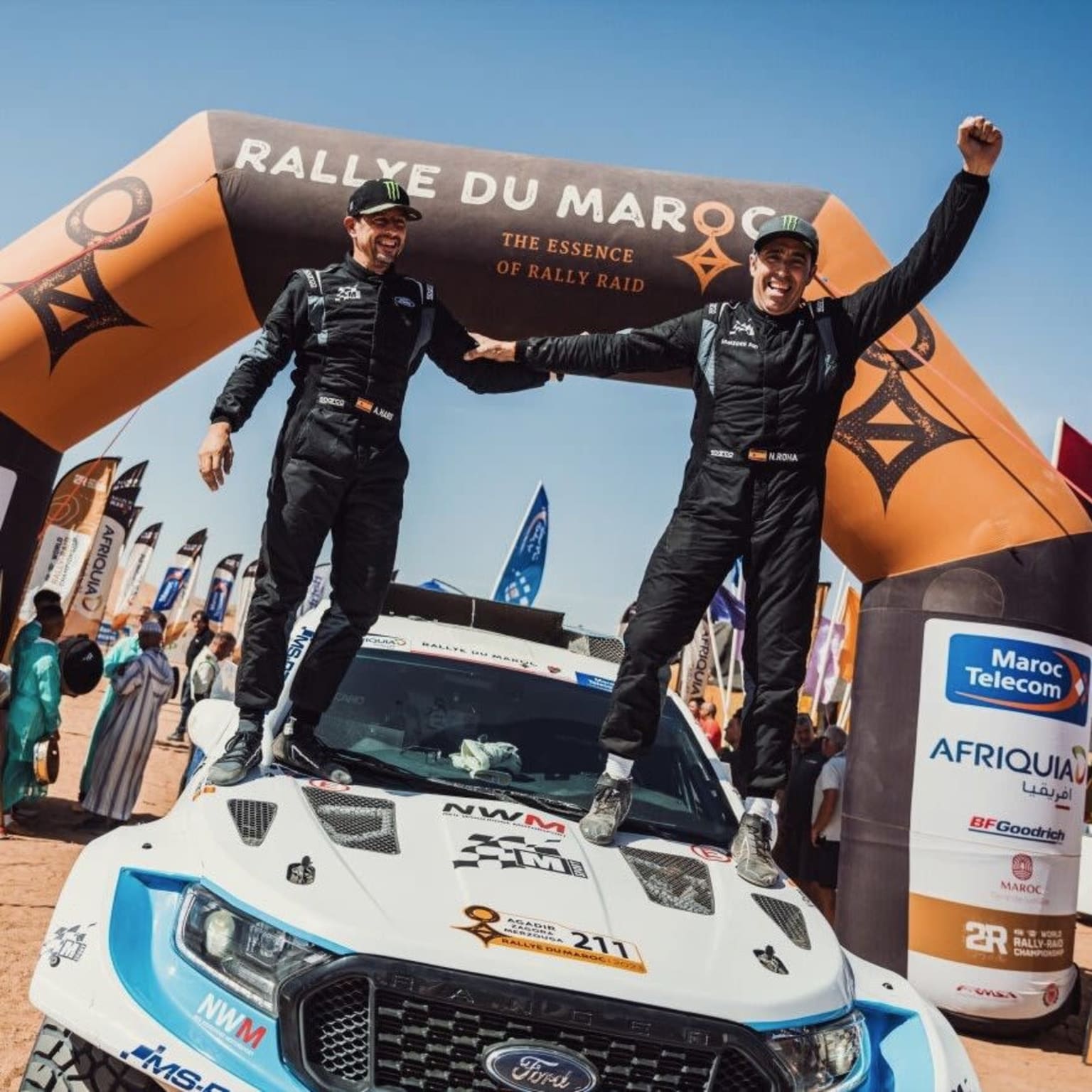 two men standing on top of a car in front of a race finish line with their hands in the air