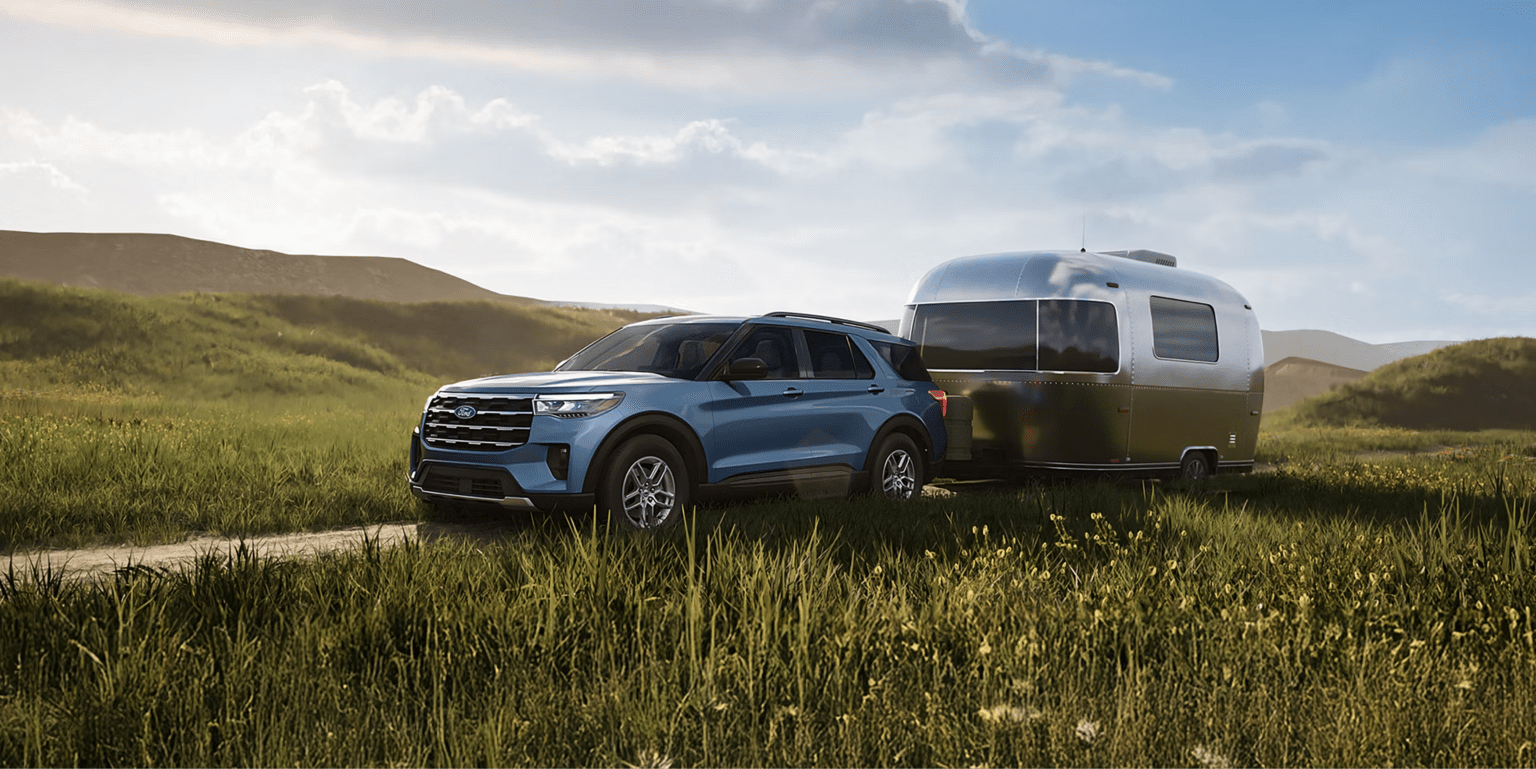 A blue SUV towing a silver travel trailer in a grassy field with rolling hills and a cloudy sky.