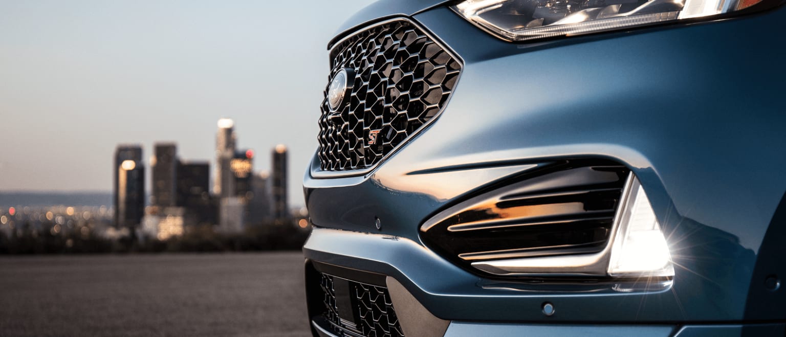 A sleek, modern car with a distinctive grille and headlights is parked on a road, with a cityscape of tall buildings visible in the background.
