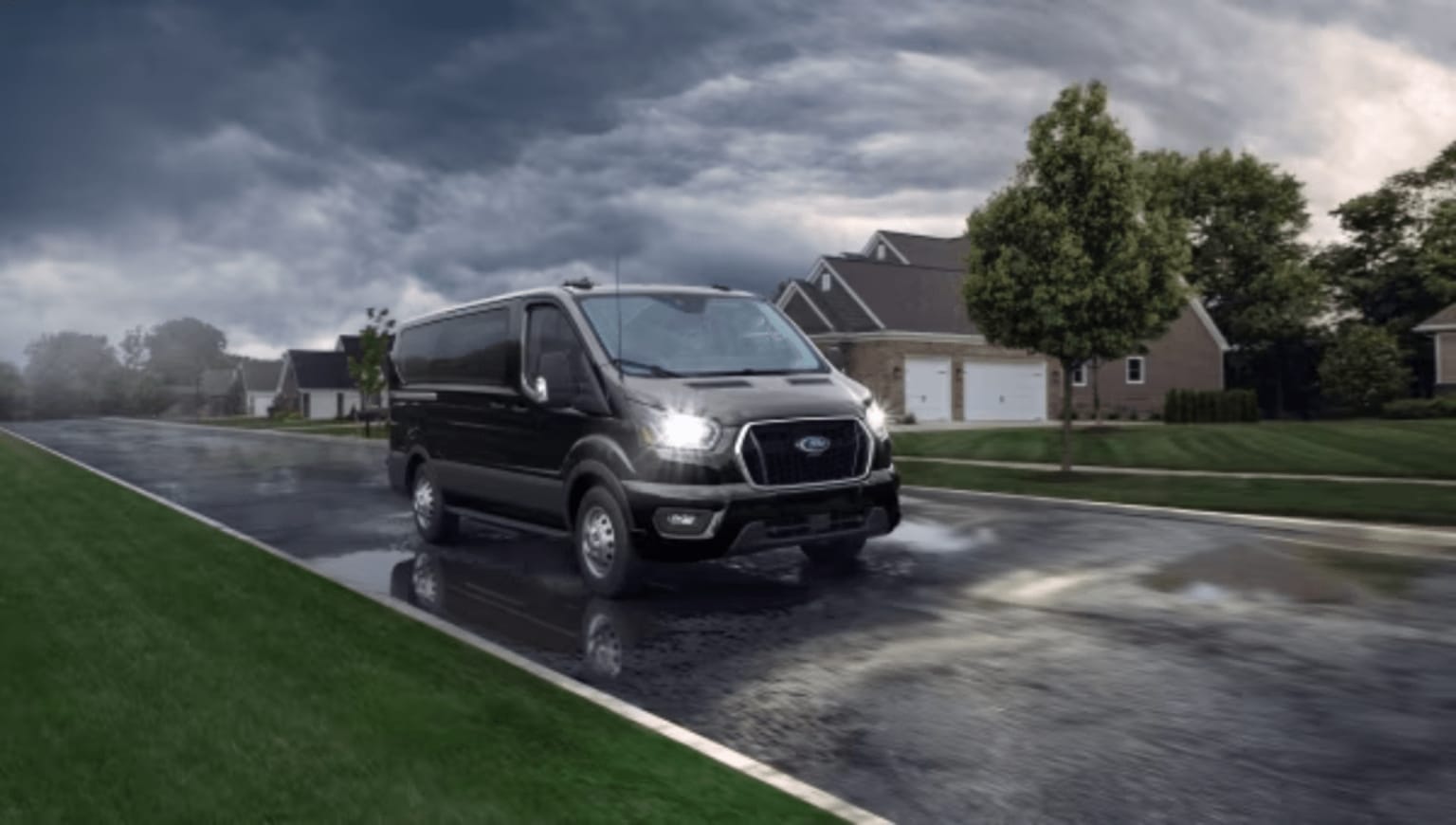 A black van driving on a wet suburban street under a stormy sky with houses in the background.