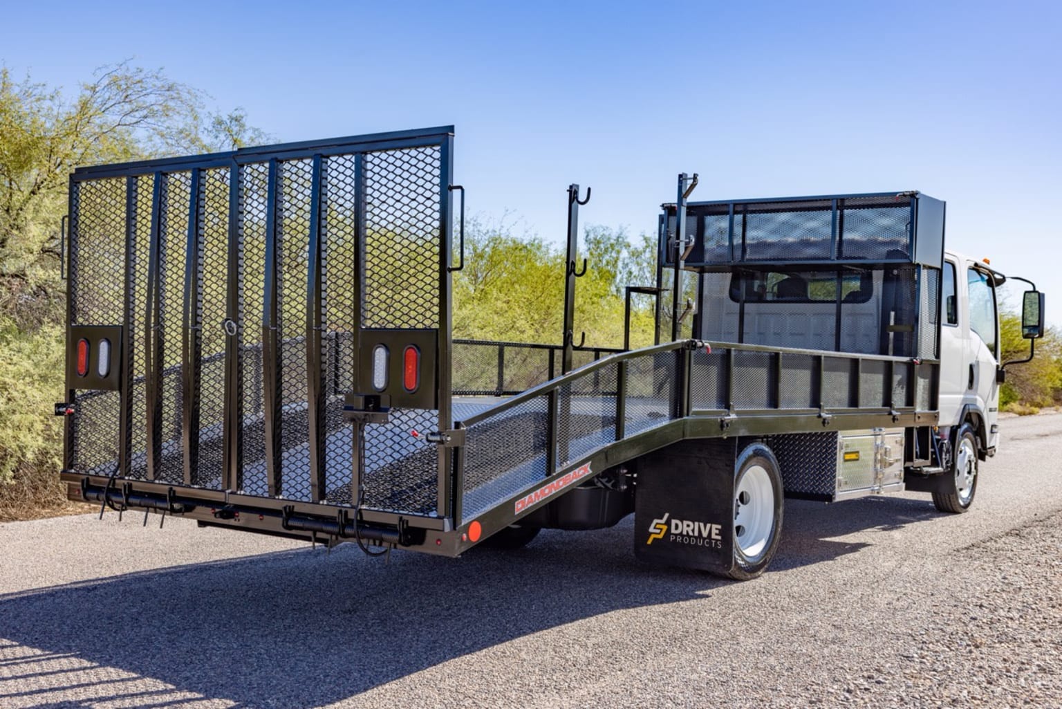 A flatbed truck with a ramp and metal railing on a paved road in front of a desert landscape with sparse vegetation under a clear blue sky.