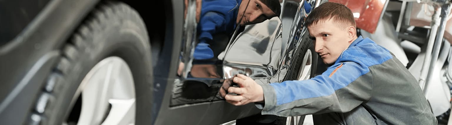A mechanic in a blue shirt is working under a vehicle, with other people and vehicles in the background.
