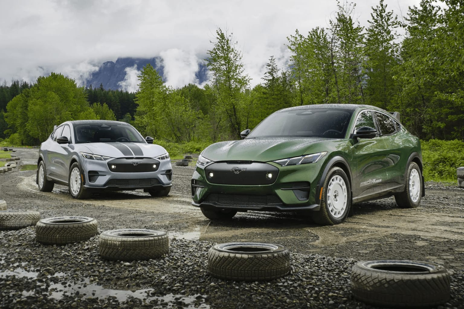 Two modern SUVs on a dirt road in a forested area with mountains in the background.