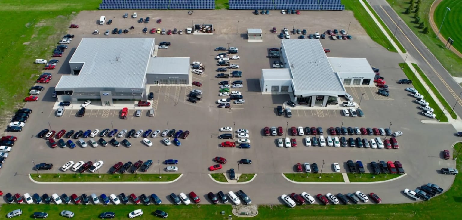 Cars parked in rows in front of a modern car dealership with solar panels on the roof in the background.