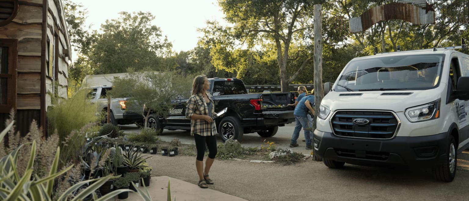 A person walking on a path surrounded by trees and parked vehicles, including a large truck and a van, in an outdoor setting.