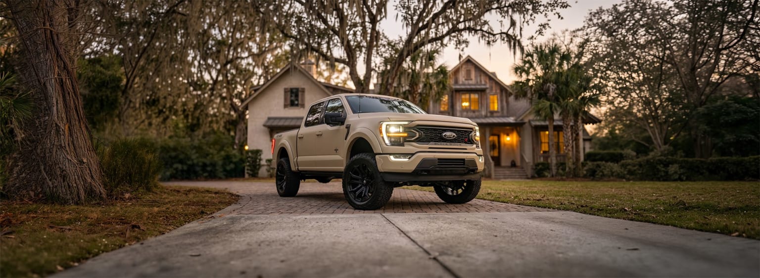 White pickup truck with off-road modifications parked in front of a two-story house surrounded by trees.