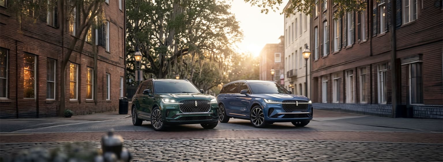 Two SUVs are parked on a cobblestone street in front of brick buildings with trees and a warm sunset in the background.