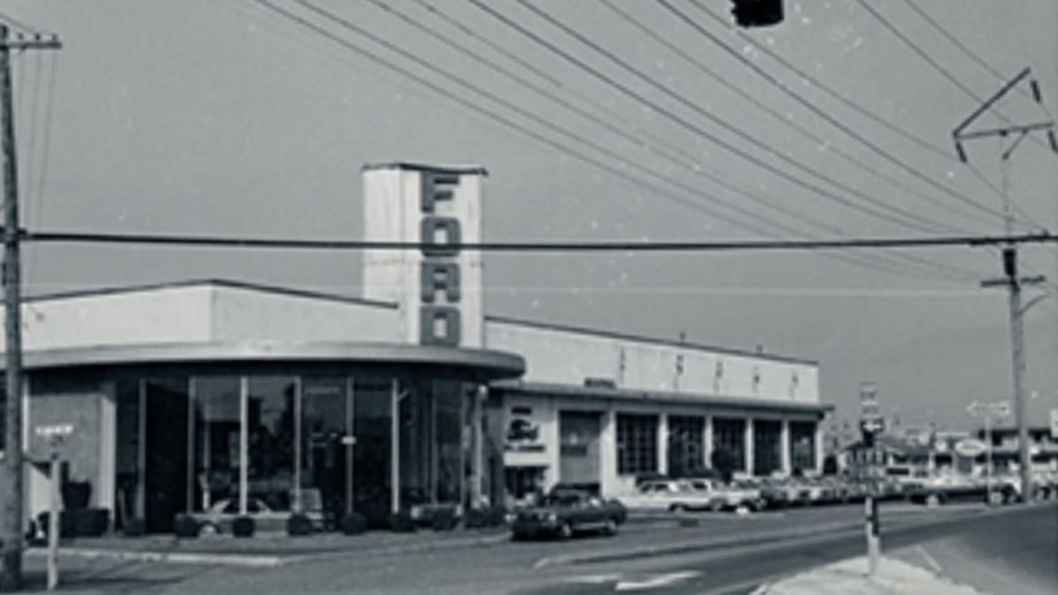 A black and white photograph of a row of storefronts with a large illuminated sign for a business called "Forno" in the foreground, with power lines and poles visible in the background.