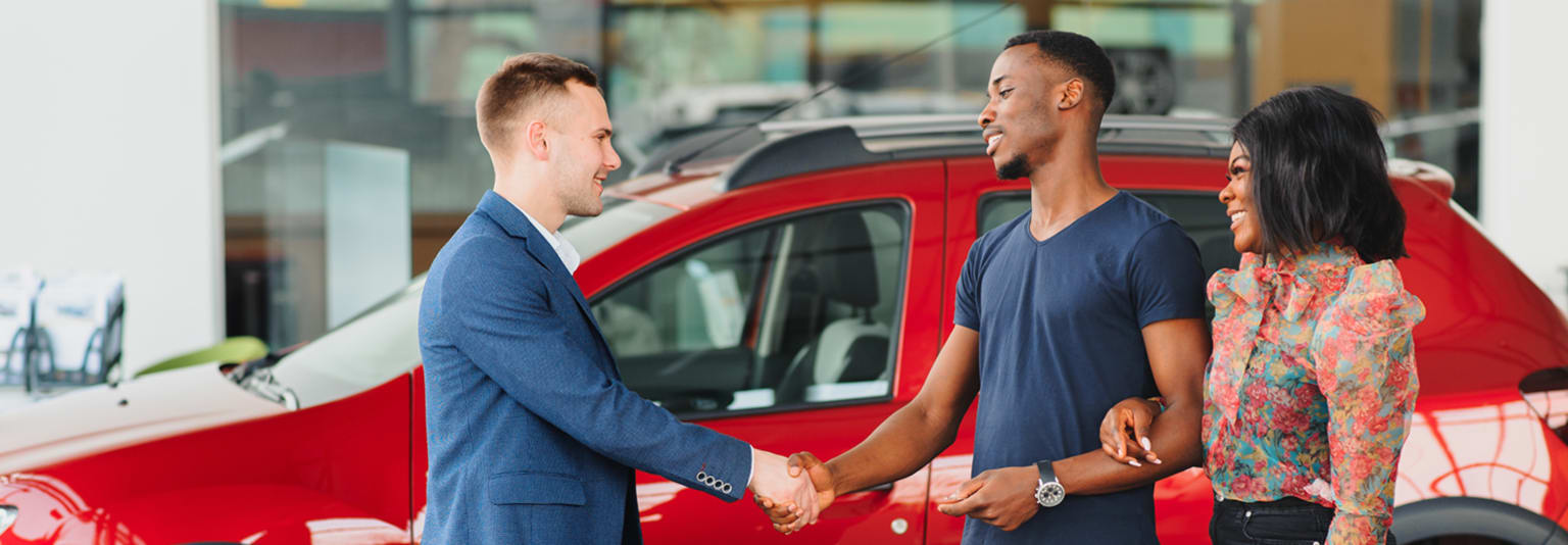 Two individuals in casual attire shake hands in front of a red car, with a woman standing nearby.