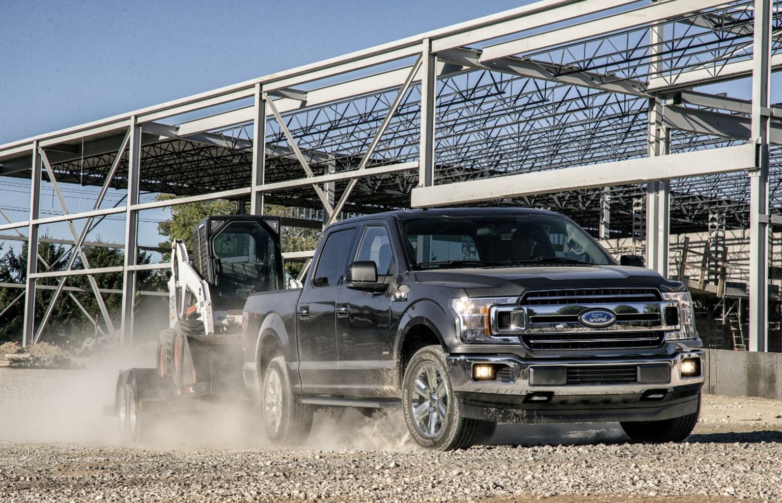 a black truck driving down a dirt road next to a metal structure with a tractor attached to the back of it