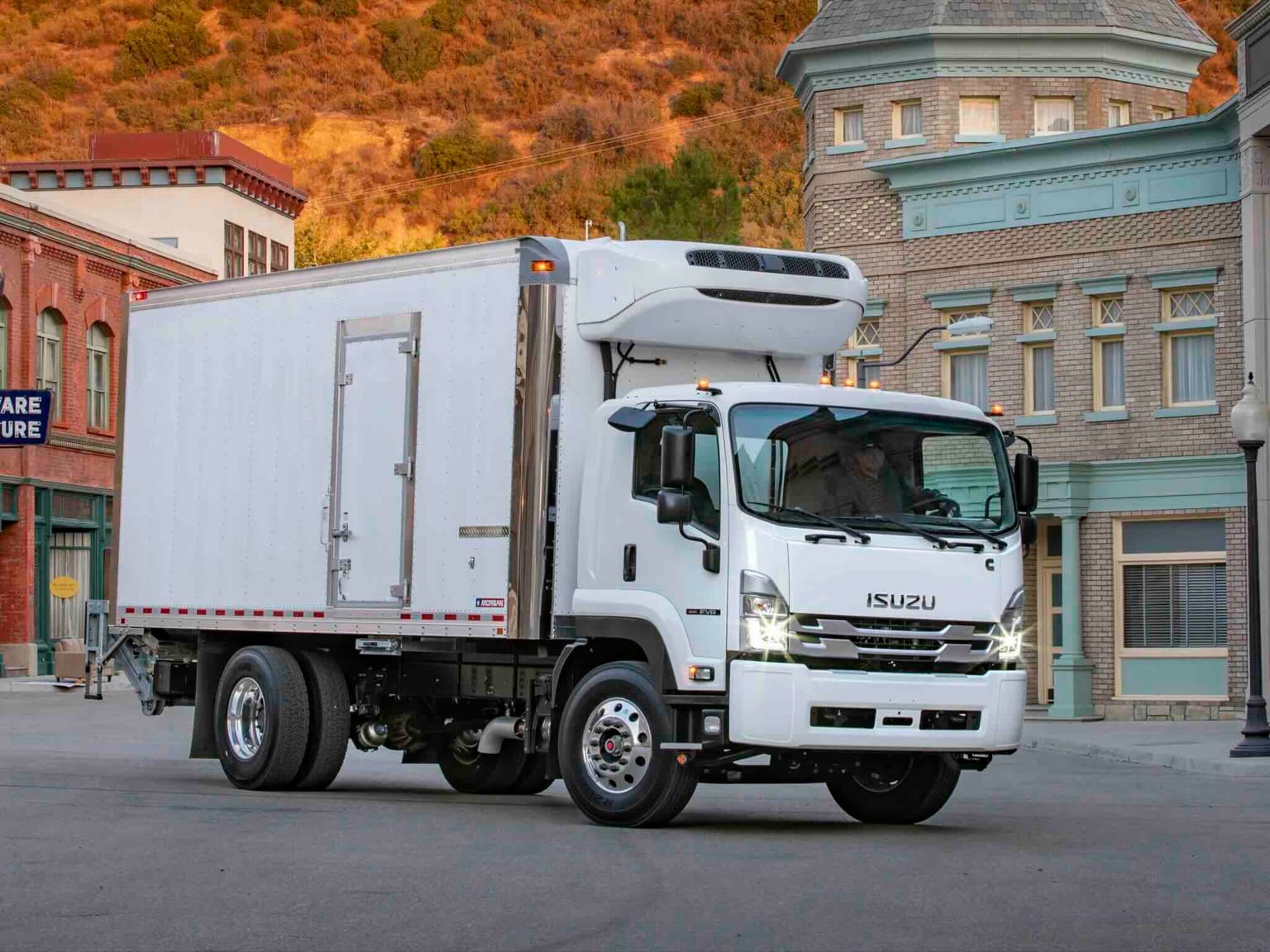 Refrigerated truck parked on a street in front of old-fashioned buildings with a hillside in the background.