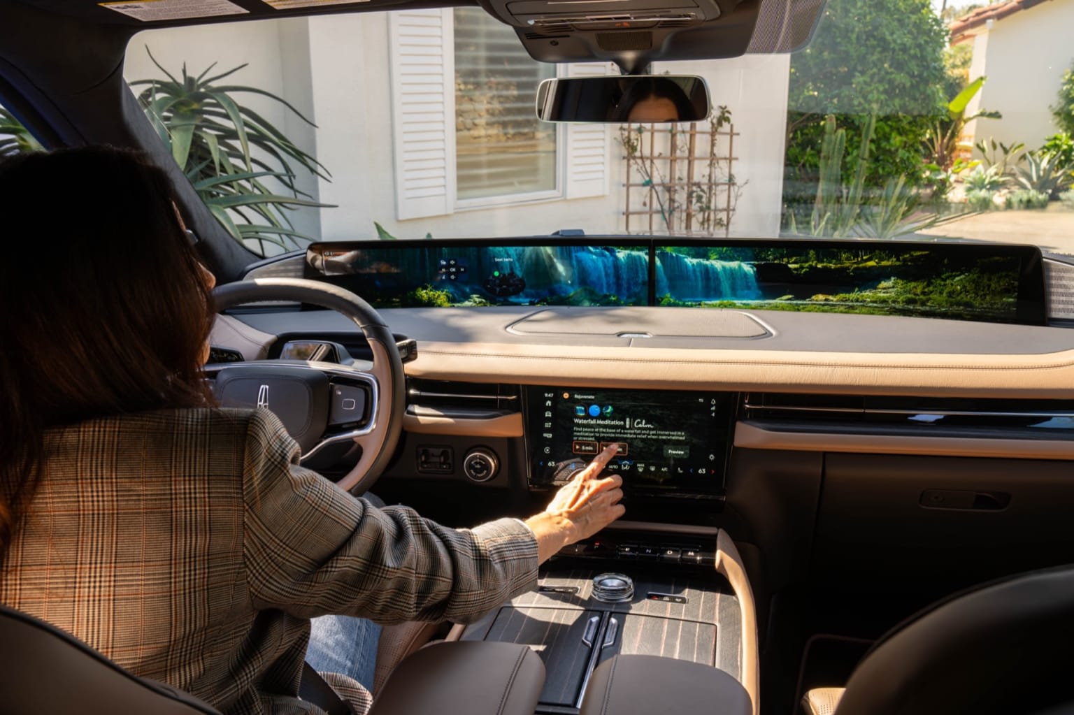The image displays the interior of a vehicle, with a person seated in the driver's seat engaging with the dashboard and controls. A scenic view, possibly of a waterfall or other natural landscape, is visible through the windshield in the background.
