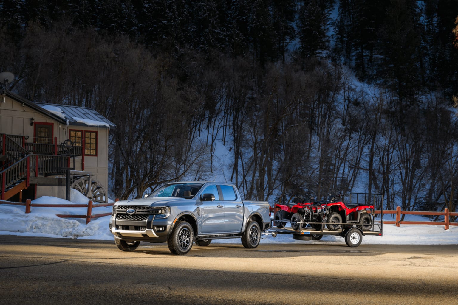 A large silver pickup truck towing a red trailer loaded with equipment, parked in front of a snowy forest and a small wooden building.