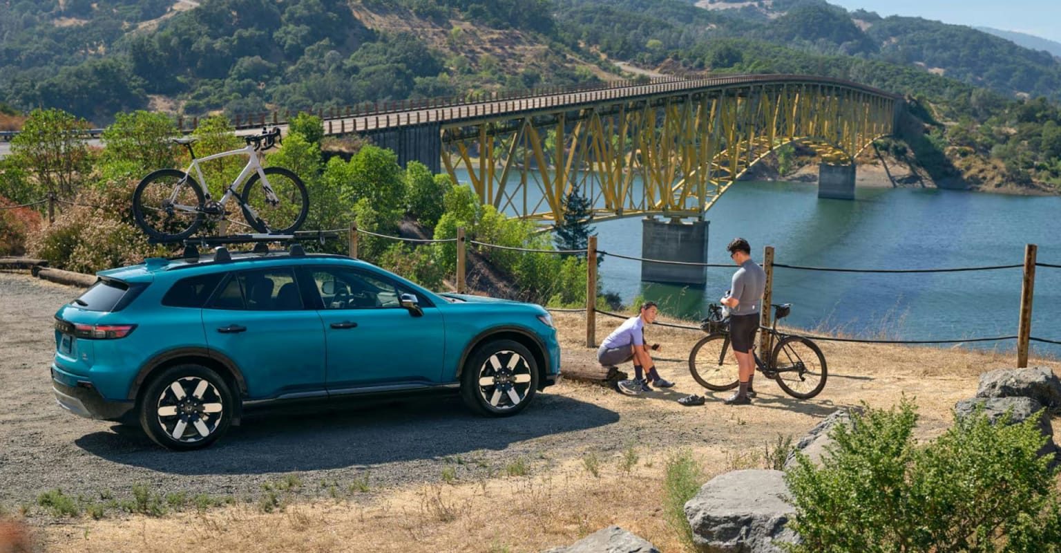 Teal SUV with bikes on its roof parked on a dirt road with two people and their bikes beside it, a large yellow bridge crossing a body of water in the background.