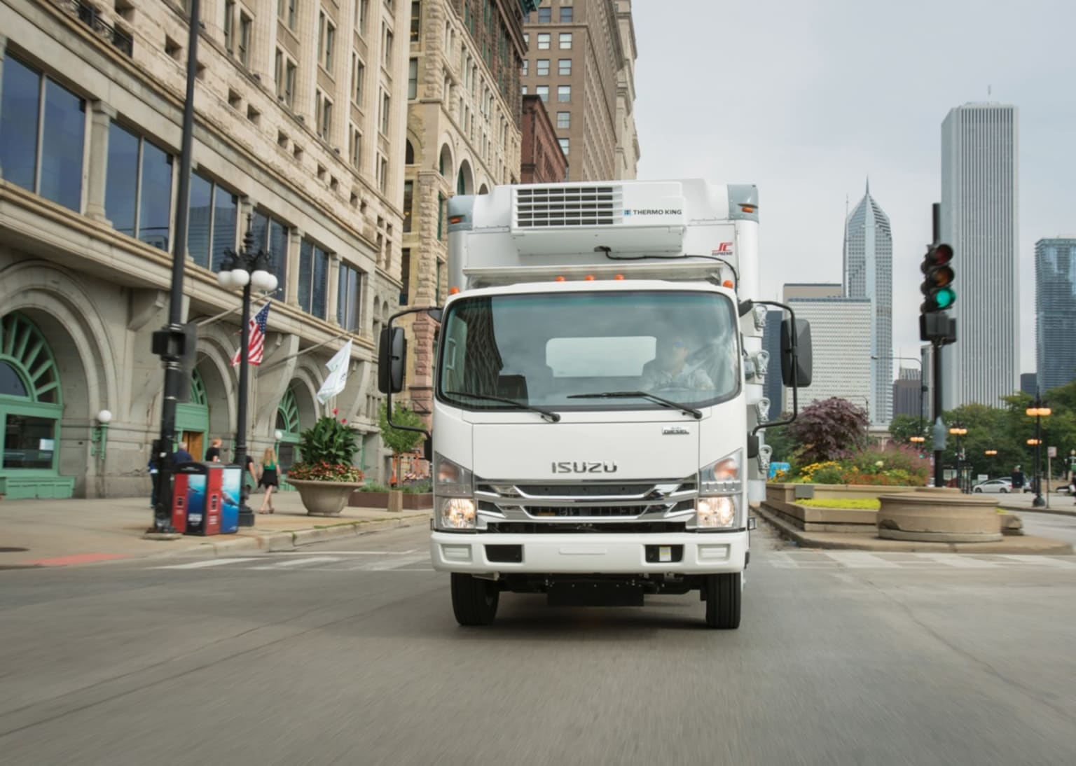 A white refrigerated truck drives down a city street with tall buildings and traffic lights in the background.