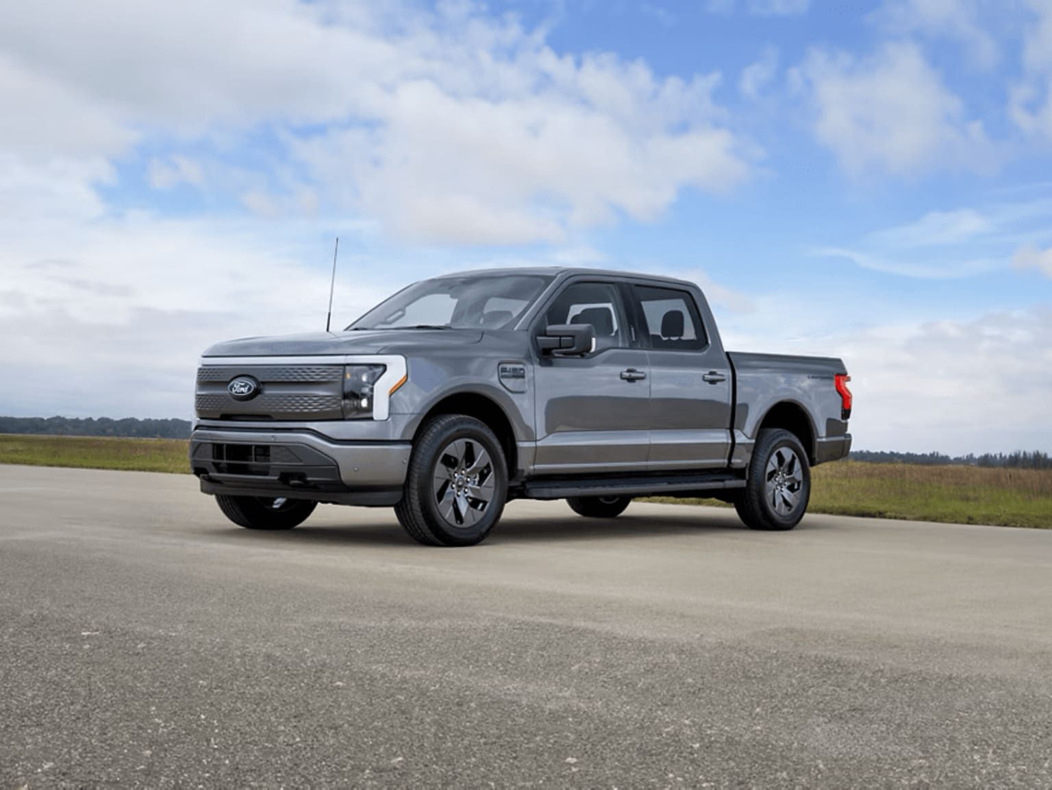 A silver pickup truck parked on a paved road with a grassy field and cloudy blue sky in the background.