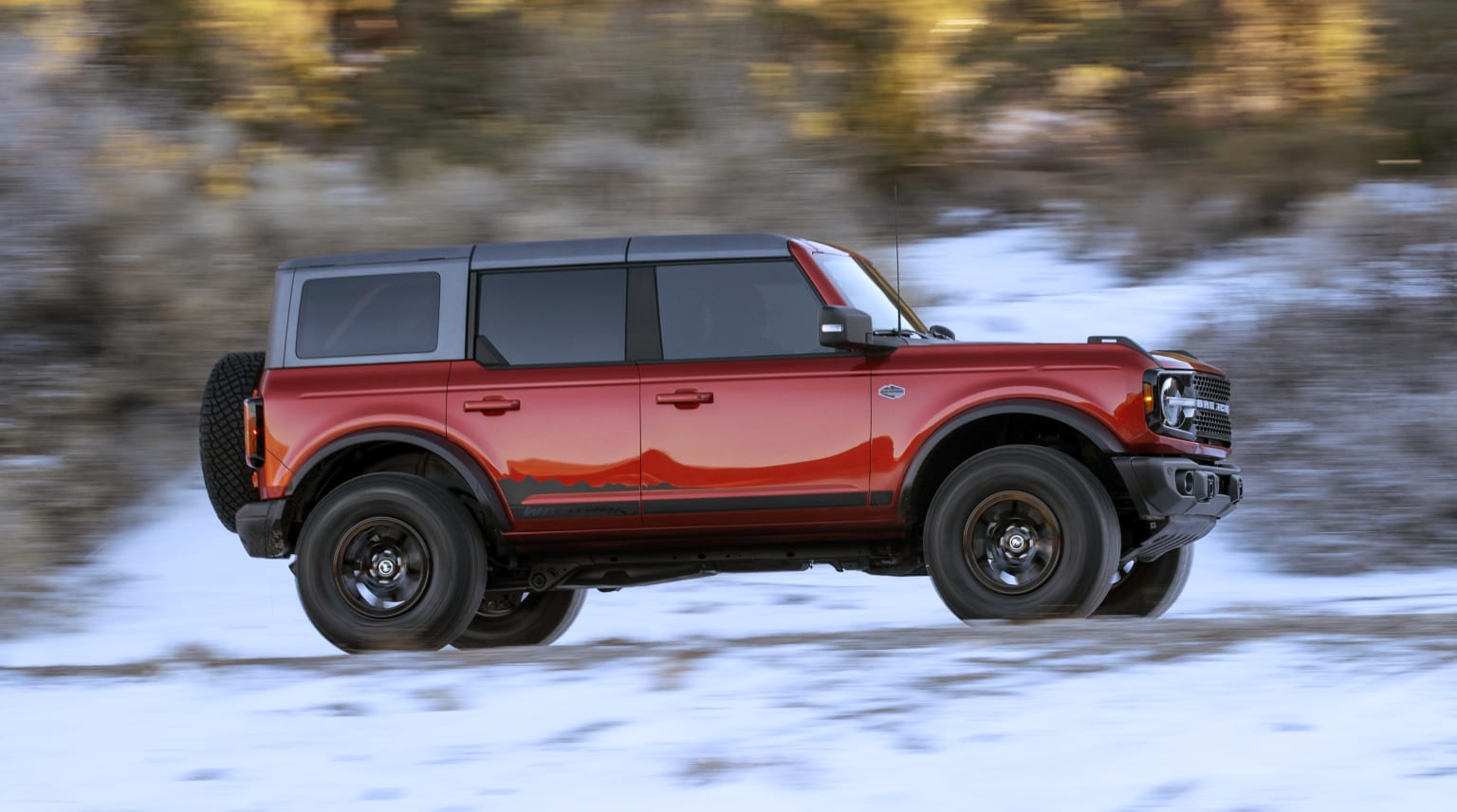 a red pick up truck driving down a road in front of a wooded area with snow on the ground