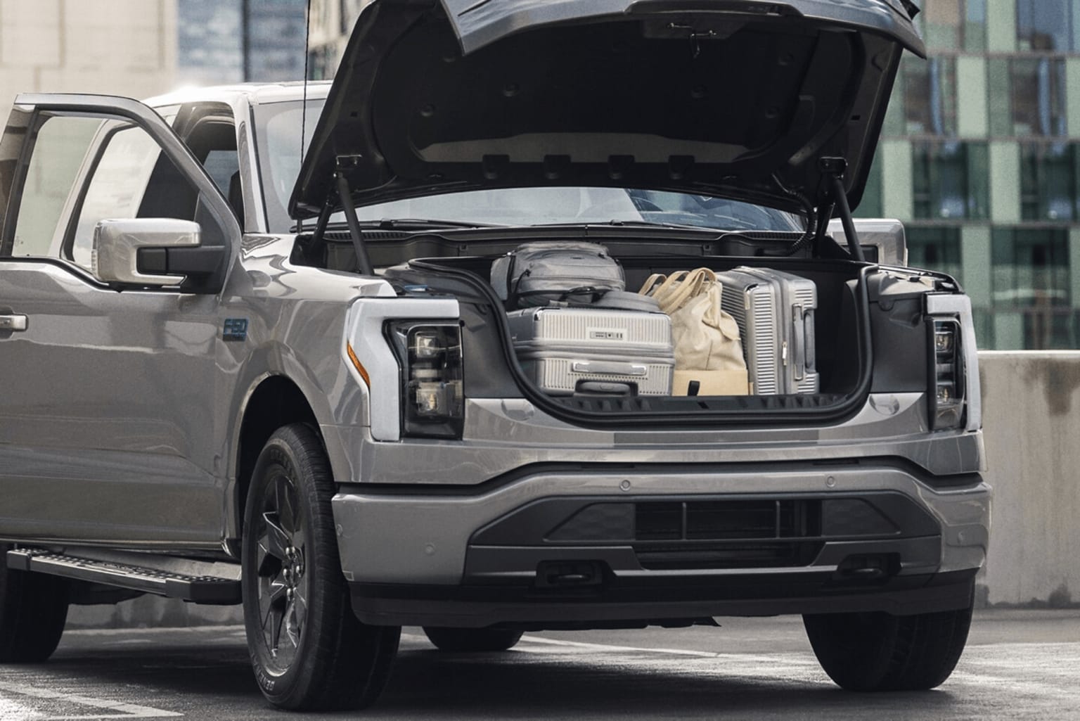 A silver SUV with its trunk open, revealing various items inside, is parked in front of a modern glass building.