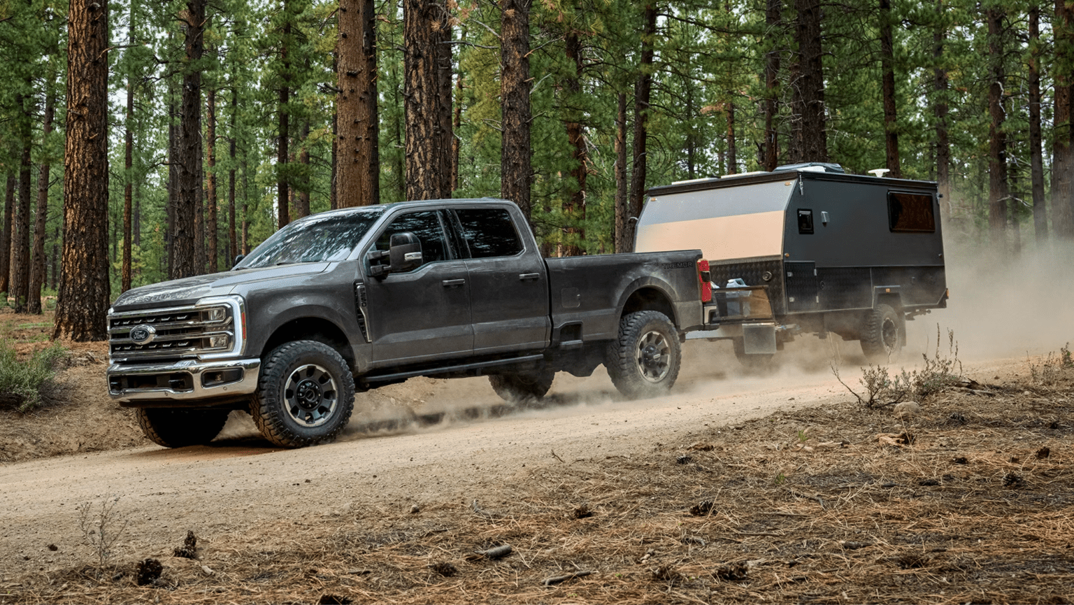 A grey pickup truck towing a camping trailer on a dusty dirt road in a pine forest.