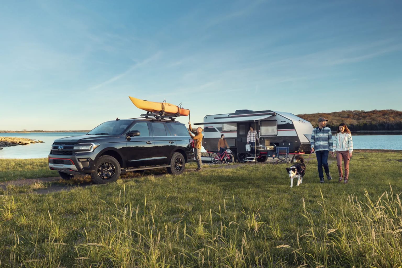 A large black SUV with kayaks on the roof parked in a grassy field near a body of water, with a group of people and a dog standing nearby an RV trailer.
