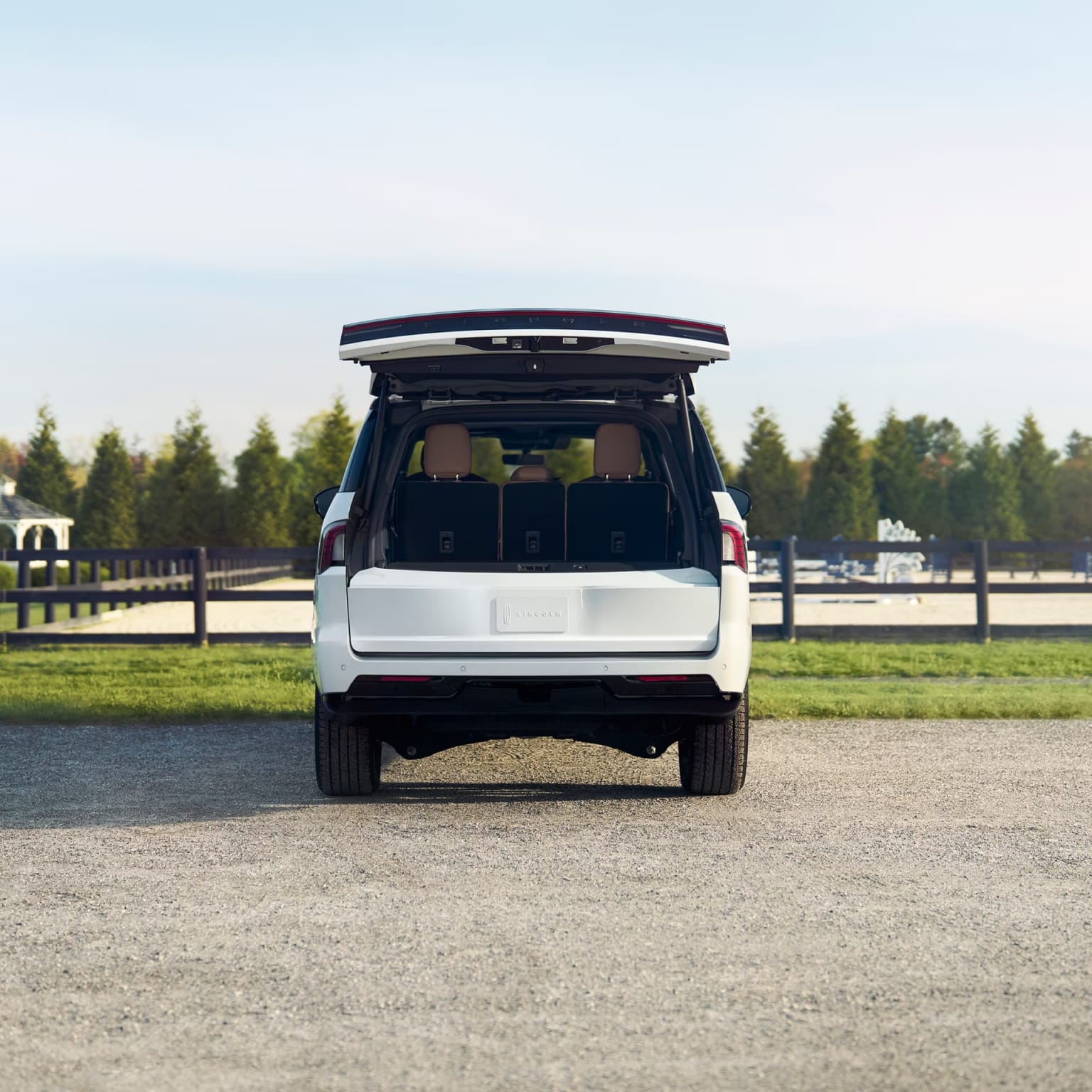 White SUV with open rear hatch parked on a gravel driveway, surrounded by grassy field and line of trees.