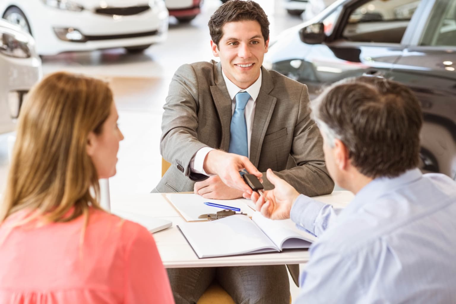 A man in a suit smiling and engaged in a conversation with two individuals at a desk, potentially in a business setting like a car dealership.