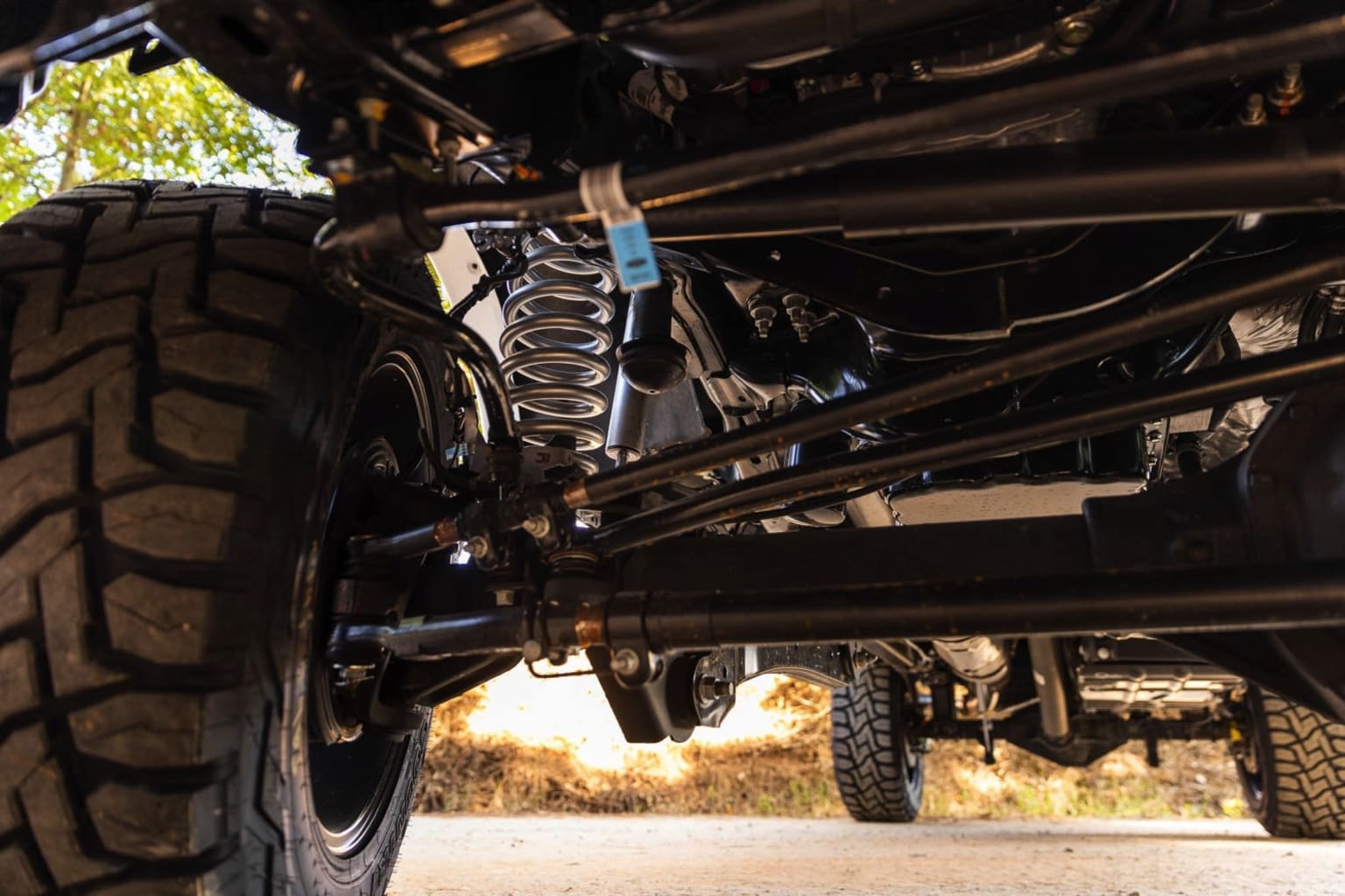 Undercarriage of an off-road vehicle with large tires, suspension components, and detailed mechanical structure against a blurred natural background.