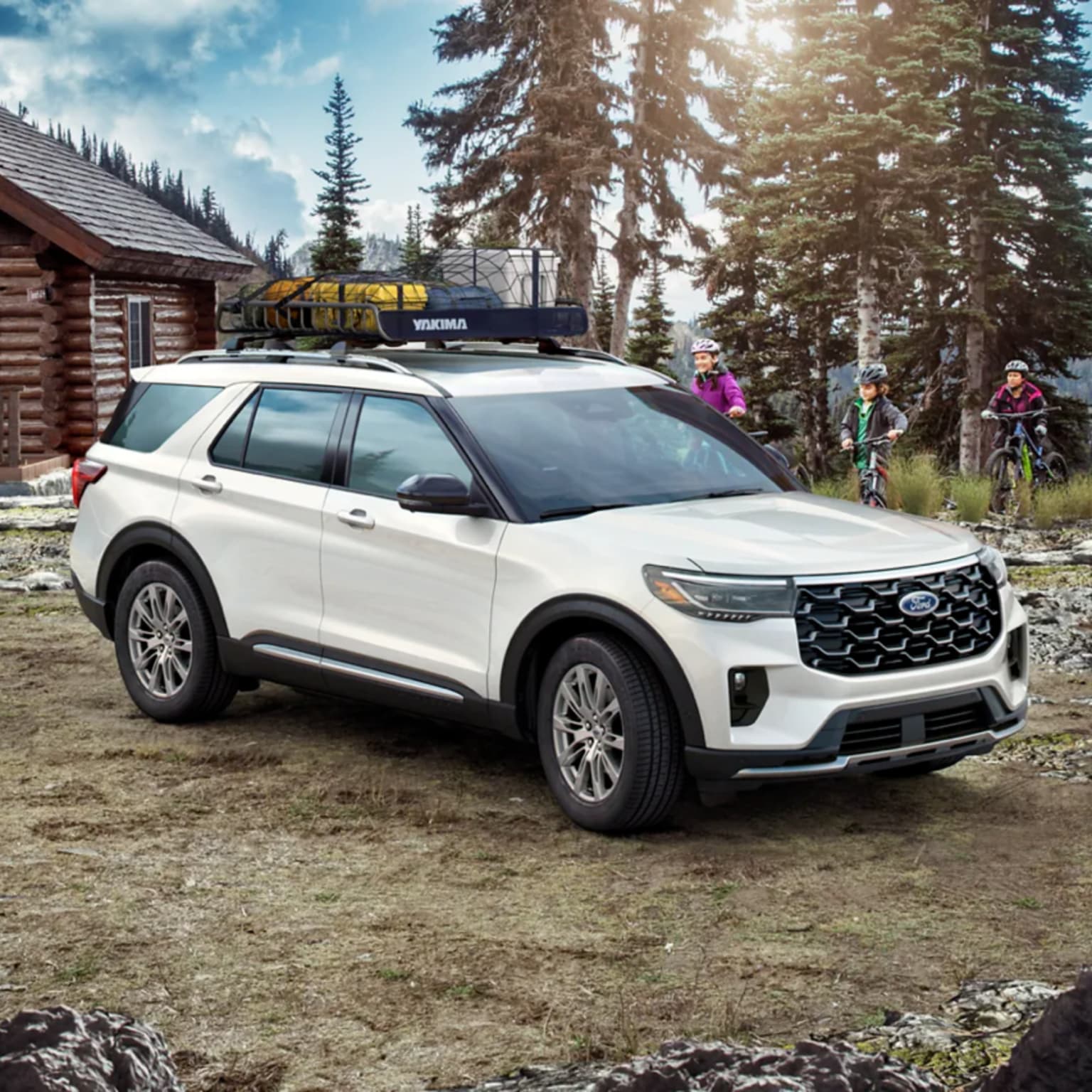 A white SUV with a roof rack is parked on a dirt path in front of a log cabin, with two people on bicycles in the wooded background.