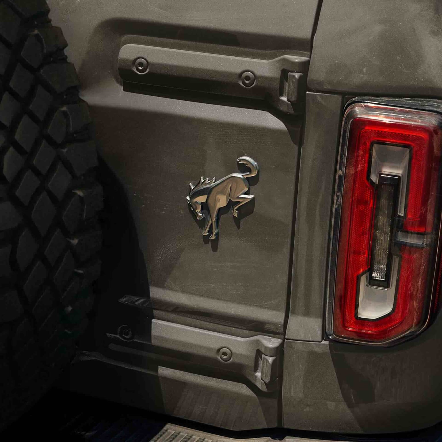 Close-up of a dark gray vehicle's rear featuring a chrome bronco emblem and taillight in the foreground, with a spare tire and textured paneling in the background.