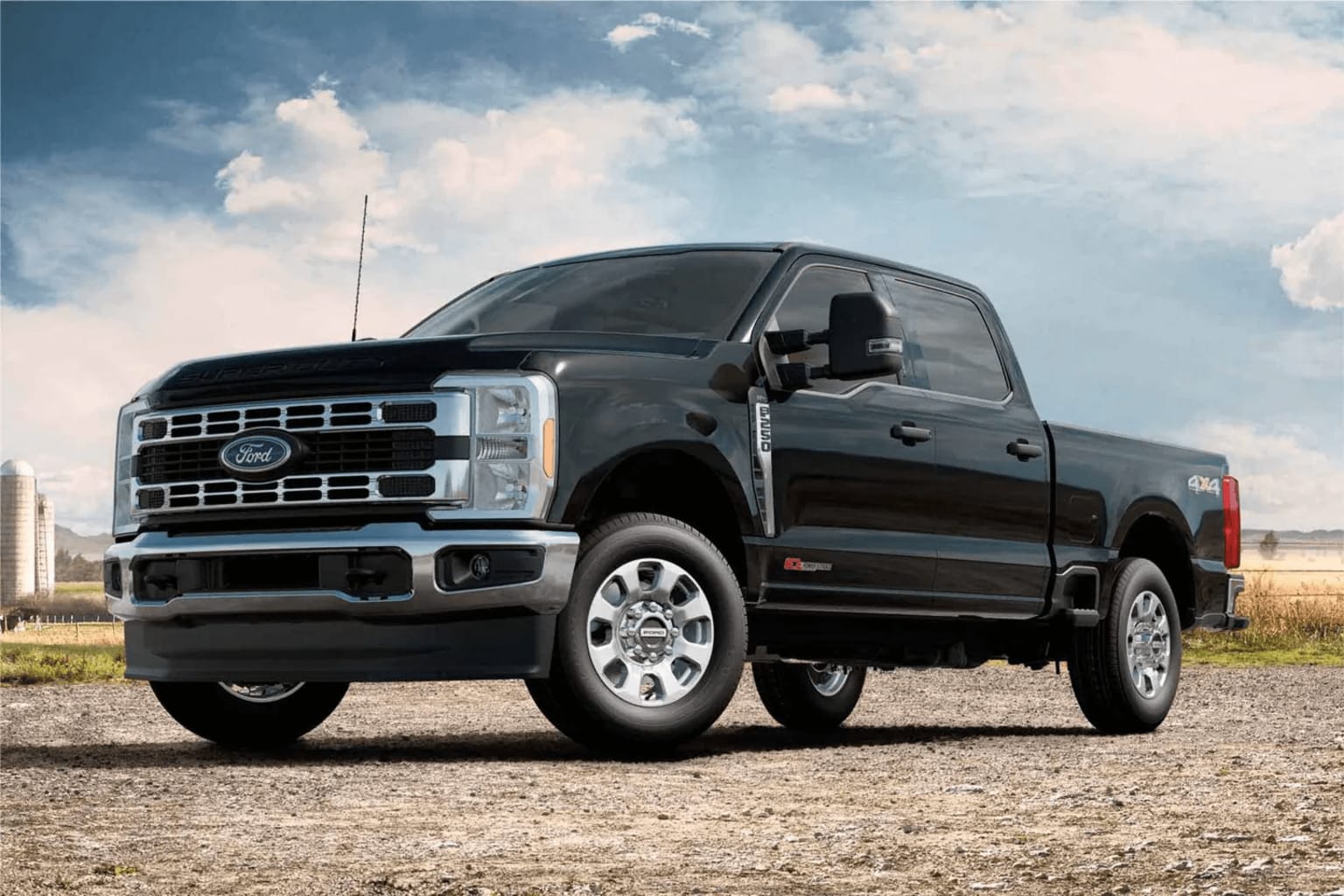A large, black pickup truck stands prominently in the foreground, set against a backdrop of a cloudy sky and a rural landscape.