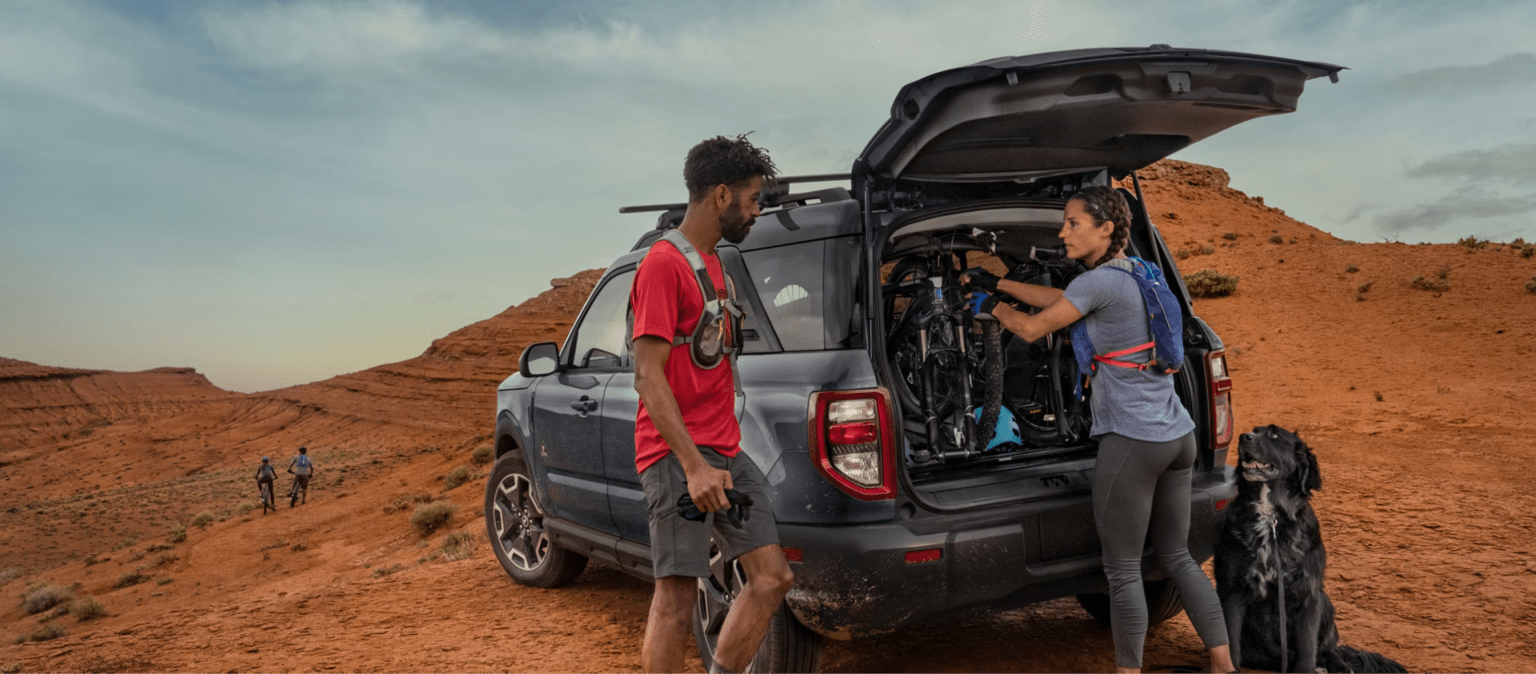 A man and woman load bicycles into a car with a dog nearby, set against a backdrop of red rock formations under a cloudy sky.