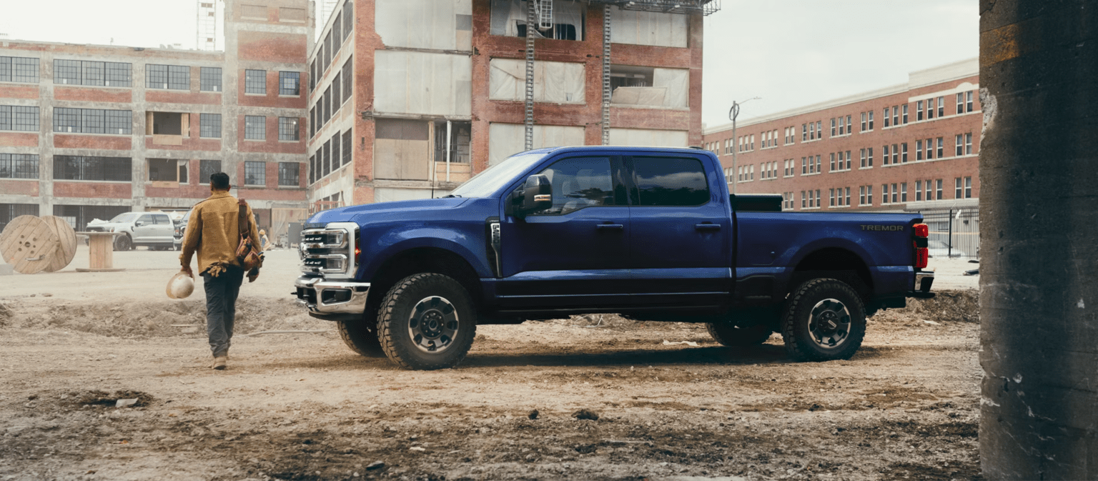 A man walks away from a blue pickup truck in a dusty industrial area with brick buildings in the background.
